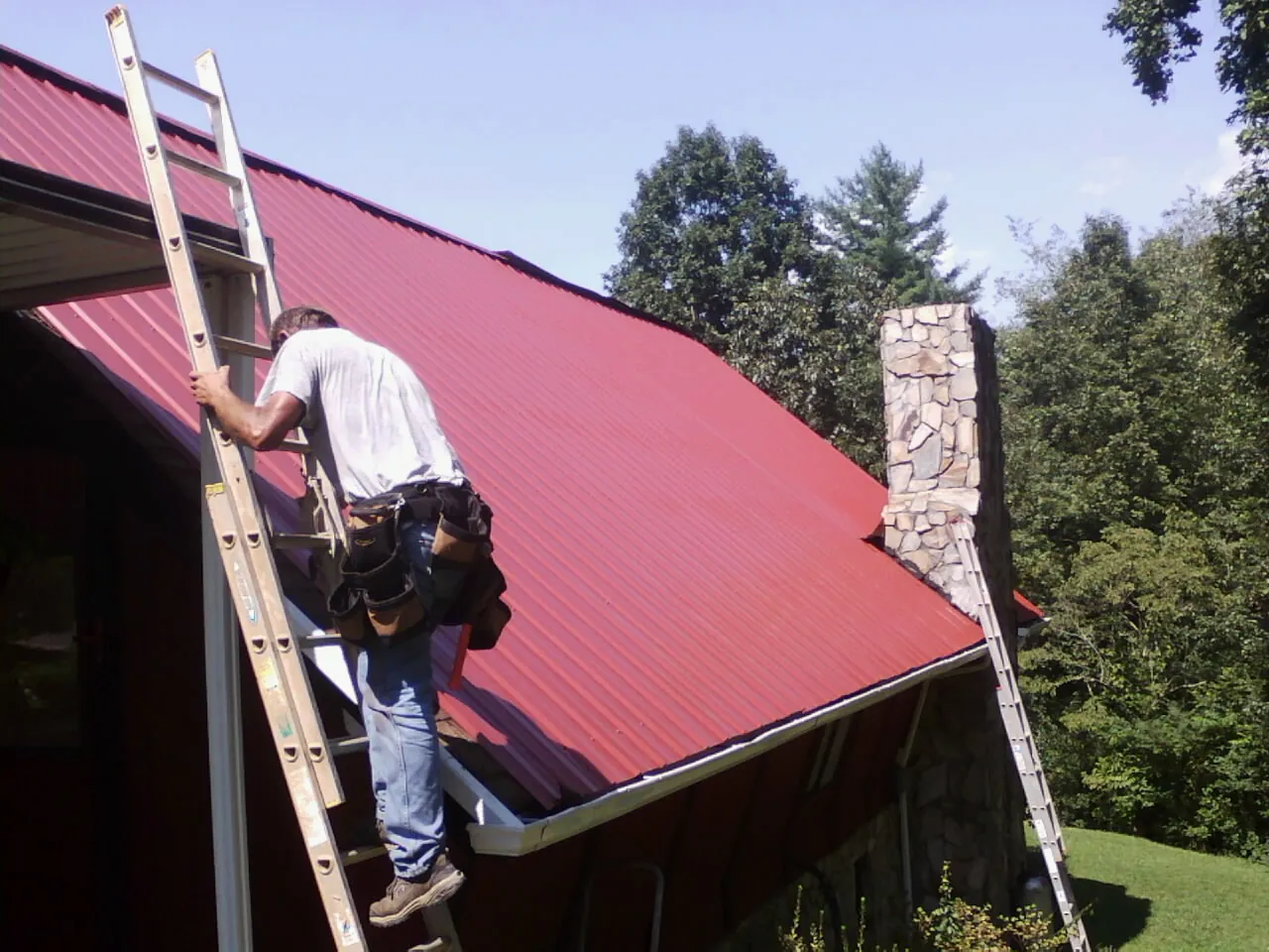 Roofer on a ladder beside a red metal roof and stone chimney — Soffit & Fascia Repair by Roofing Brothers in Boonton